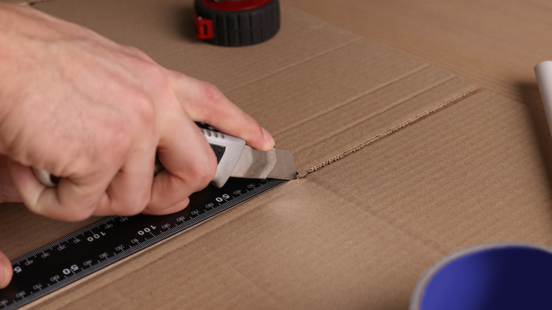 A light-skinned hand cuts cardboard with a utility knife.