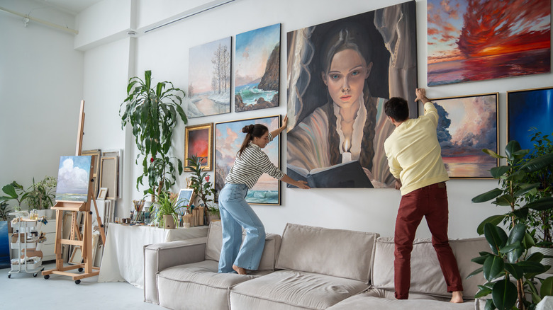 A couple hangs a large painting in the center of a gallery wall in their living room.