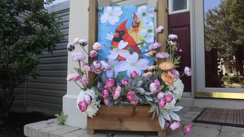 A wooden stand with a cardinal garden flag and flowers.