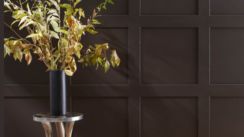 A deep brown wall with a flower vase on a stool in front of it