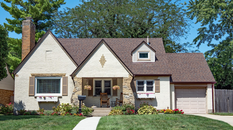 Modest, two-story, tan brick and flagstone house with large flowerbed and large background trees.