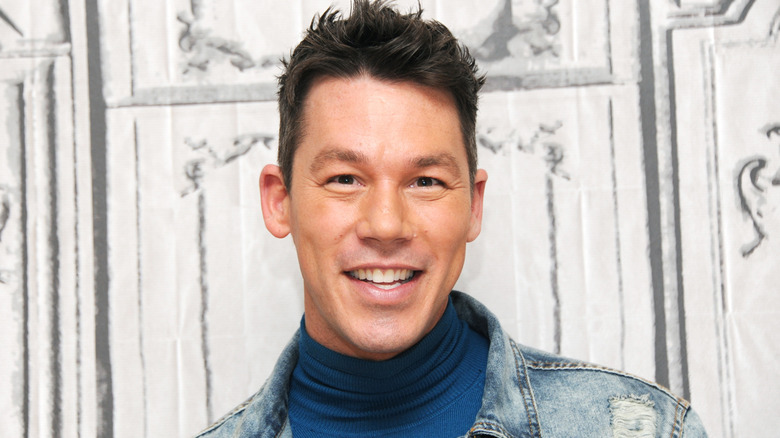 David Bromstad laughing into a microphone at the AOL Build Speaker Series, with a poster of My Lottery Dream Home in the back
