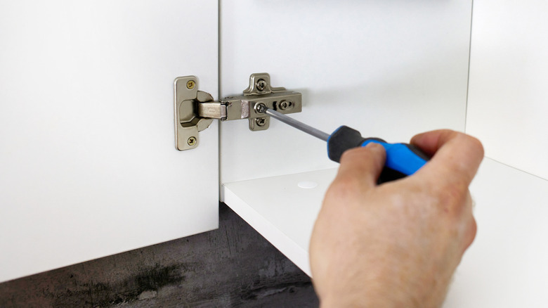Screwing a door hinge to white kitchen furniture using a screwdriver.