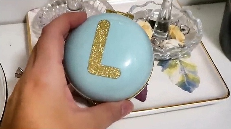 Hand holding a blue trinket box with a golden 'L' on top