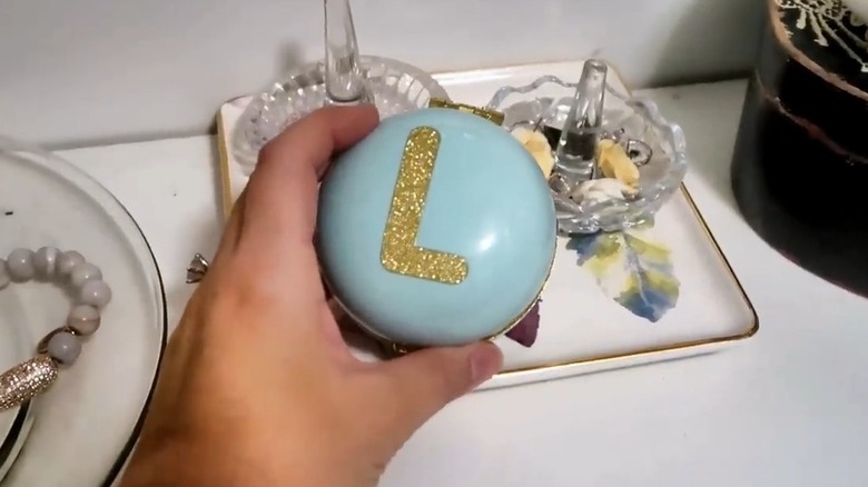 Hand holding a blue trinket box with a golden 'L' on top