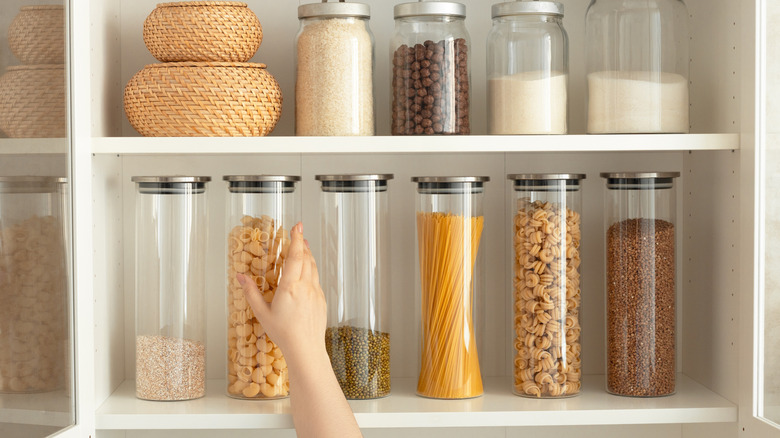 Hand takes a glass jar of pasta from a shelf in the cupboard with other jars in view