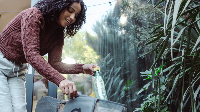 Woman disposing of a plastic water bottle into recycling bag against garden backdrop