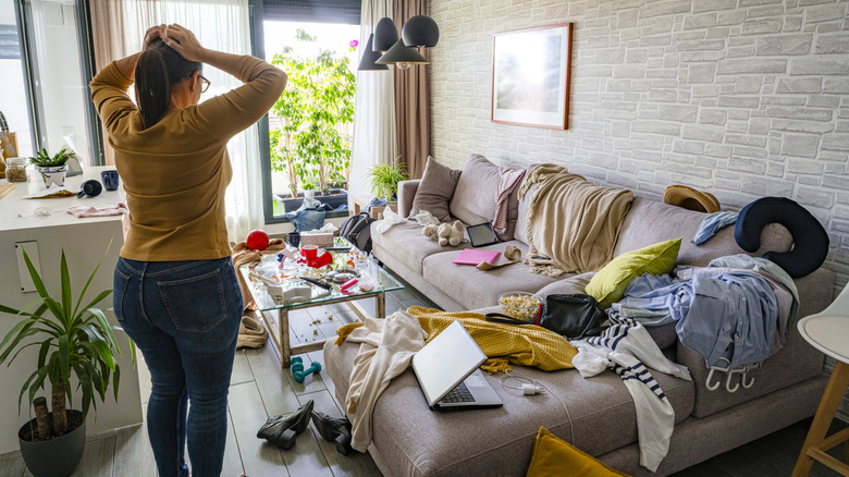 Woman standing with hands on head, overwhelmed by cluttered living room.