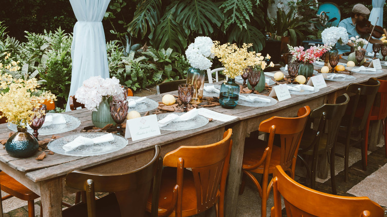 A man plays a violin at the end of a long wooden table with assorted wooden chairs