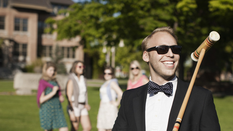 A man wearing a bowtie and sunglasses smiles while holding a croquet mallet
