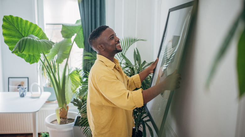 A smiling man hangs a piece of framed art.
