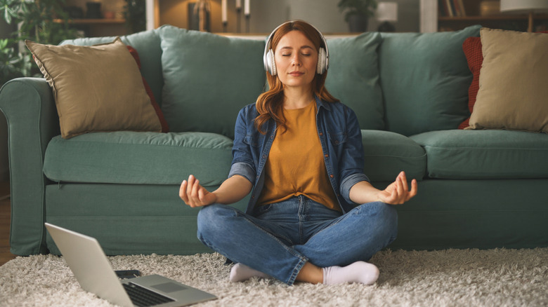 A woman with headphones on sits on a rug in front of a sofa, meditating.