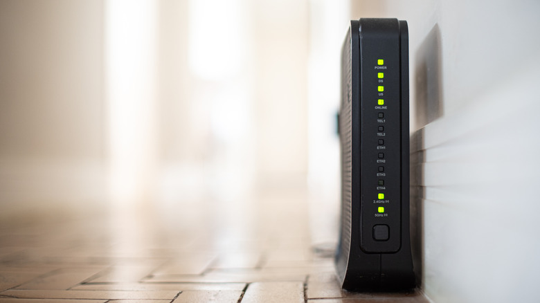 A black WiFi modem sits on the floor next to the wall in a brightly-lit room.