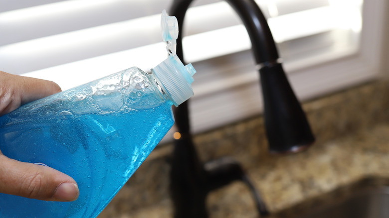 a hand holding a bottle of blue dish soap over the kitchen sink