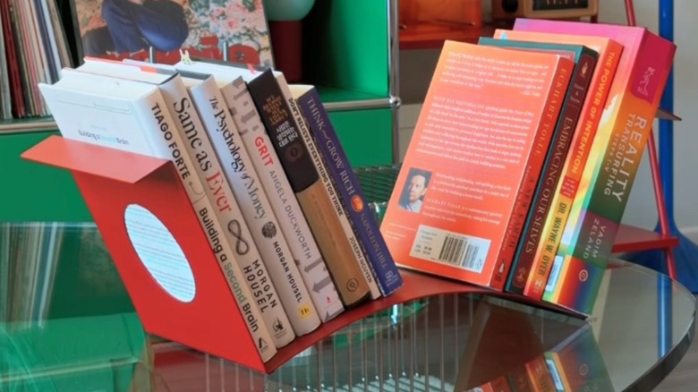 A curved red metal standalone shelf holding 11 books of various sizes and colors sits on top of a glass tabletop.
