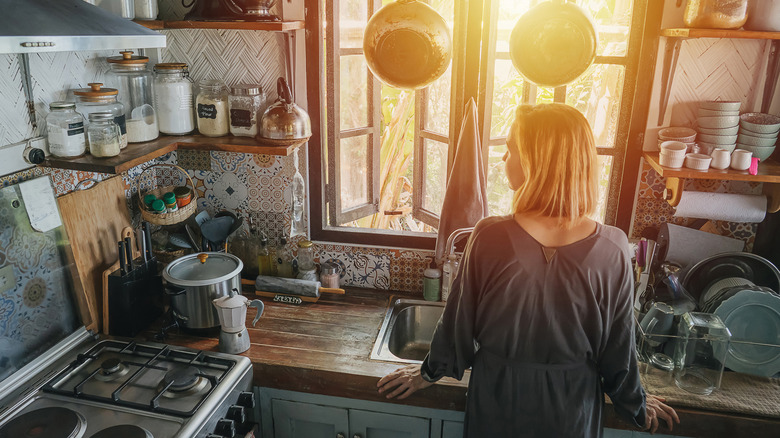 Woman standing in small kitchen that is cluttered with pots, pans, and various kitchen items.
