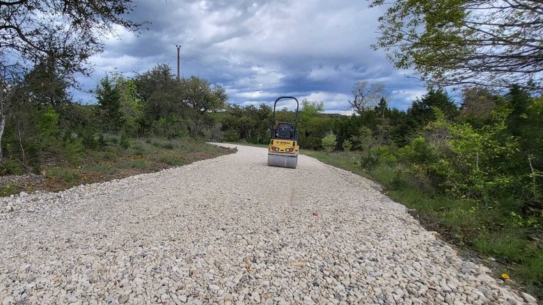 A new crushed limestone driveway with trees and bushes on either side and a yellow compactor machine in the middle of the driveway