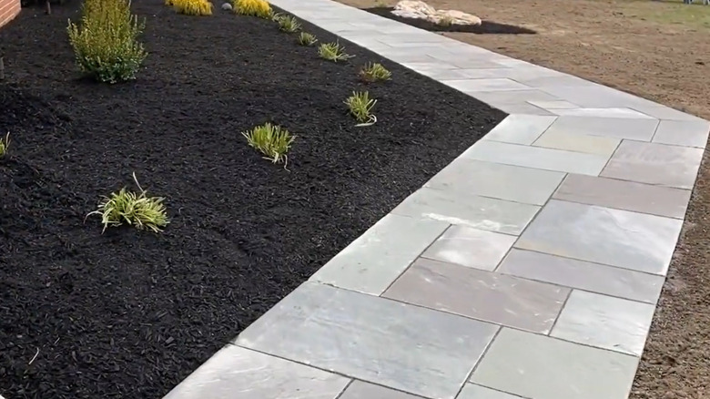 A bluestone walkway runs between a garden bed and a lawn in front of a brick house.