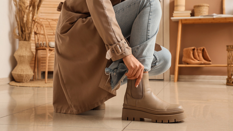A person pulls on some boots while crouching in the entryway of a home.