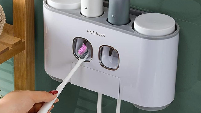 Toothpaste dispenser and holder affixed to bathroom wall with a woman's hand insterting a toothbrush into the dispenser