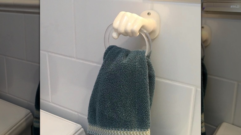 Ceramic hand holding hand towel ring with towel hanging from it, mounted on a tile wall.