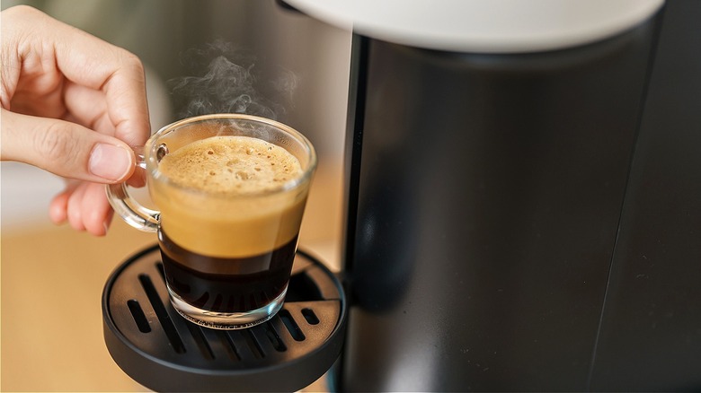 Close-up of a person's hand holding the tiny handle of a steaming cup of single-serve coffee