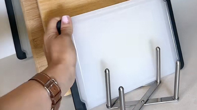 A hand puts a white plastic cutting board into an IKEA VARIERA pot lid organizer on a countertop with other cuttings boards in it.