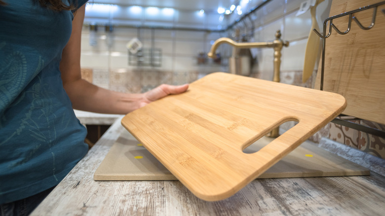 A woman picks up a wooden cutting board from a kitchen countertop cluttered with more boards, stands, and other items.