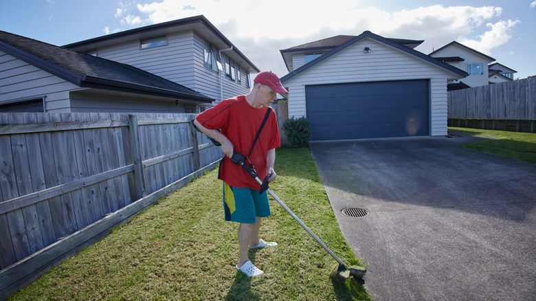 A man trims the grass at the edge of a concrete driveway using a string trimmer.