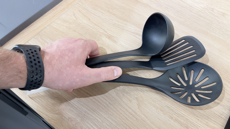 A man's hand picks up three serving utensils from a wooden countertop