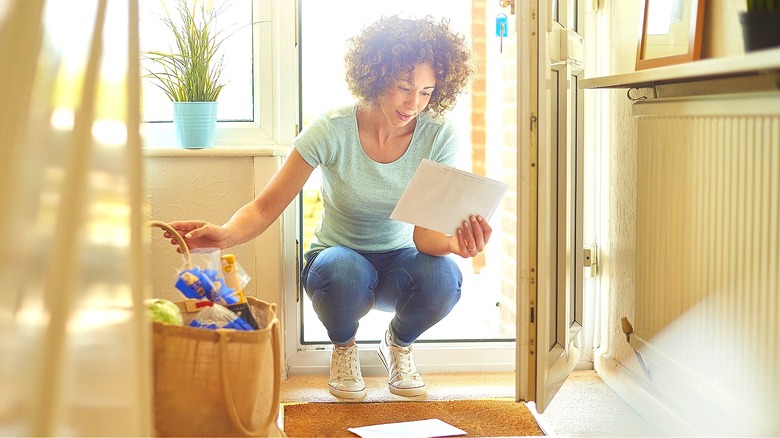 Woman looking at papers on the floor after opening the front door of home with groceries