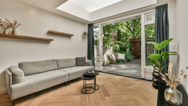 A living room with wood flooring, large glass sliding doors, drapery, and a grey couch
