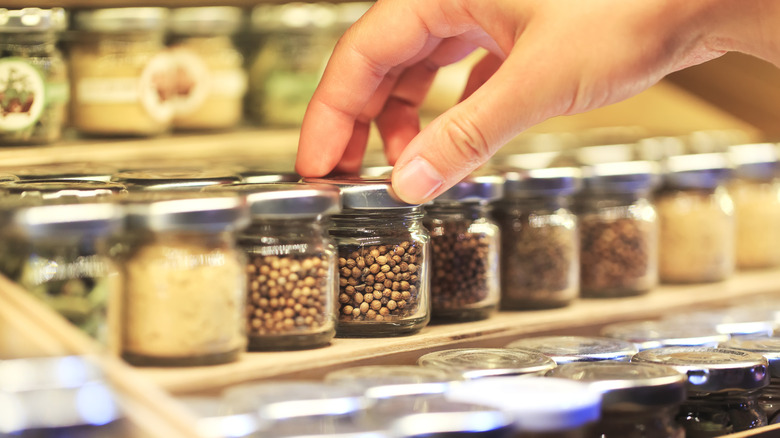 A hand reaches for a jar full of peppercorns sitting with other jars of spices on a pantry shelf.