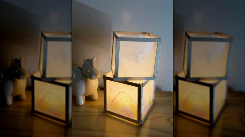 Two lanterns made of popsicle sticks and parchment paper, with children's drawings on the paper, are lit from within.