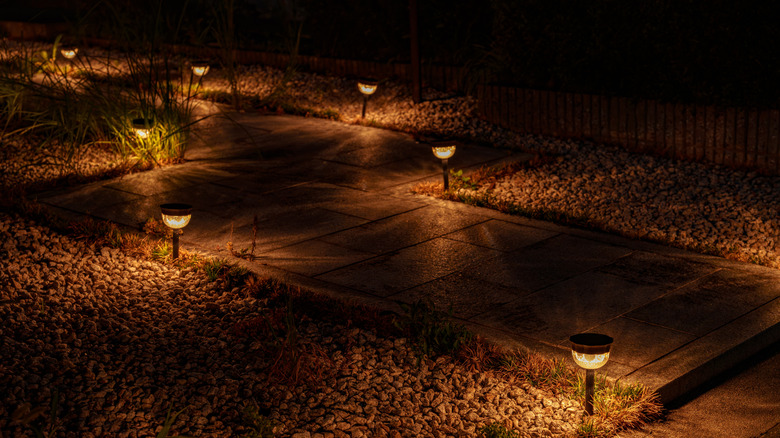 A paved garden path bordered by gravel is lit in the dark by warm yellow stake lights.