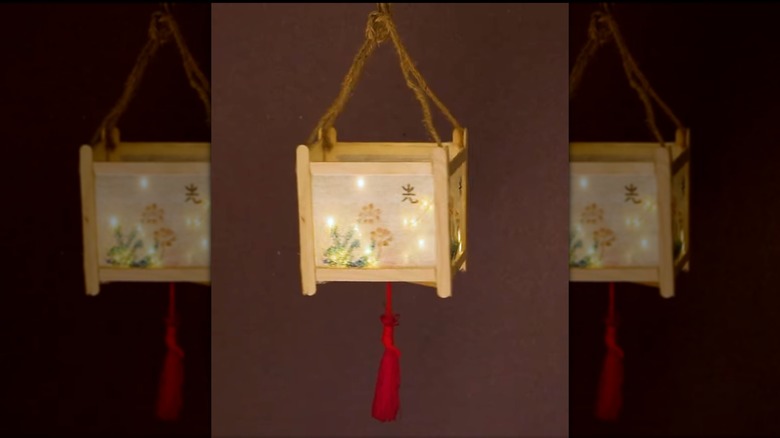 A hanging lantern made with popsicle sticks has a shade decorated with a simple flower and a red tassle underneath.