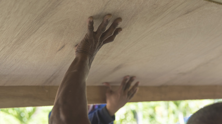 A person installing a piece of plywood on the ceiling