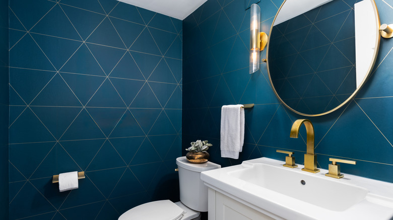 A modern bathroom with blue wallpaper, a single vanity, a round vanity mirror, and gold hardware.