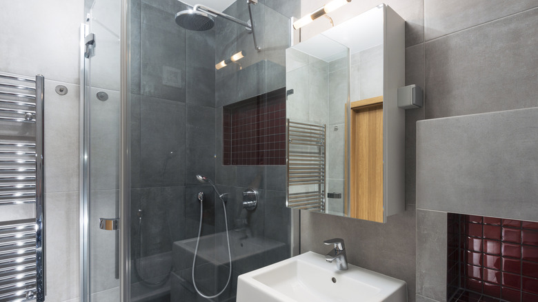 A bulky mirror fronted medicine cabinet in a modern bathroom with grey tiles, large shower with rain shower head and a chrome towel rail.