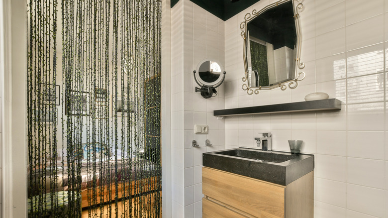 A sink with pine cabinet underneath with a floating shelf above it. There is also a large mirror attached to the wall and a magnifying mirror on an extendable arm. There are cream tils and a rafia hanging as an alternative to a door.