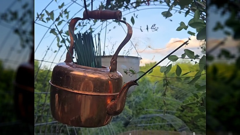 A copper kettle hanging on a hook that holds citronella sticks in the top with one burning stick in the spout.