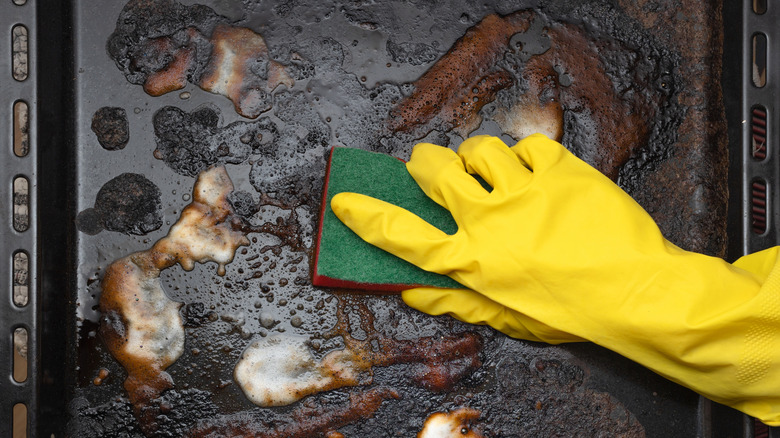 Gloved hand scrubbing a greasy baking sheet with sponge