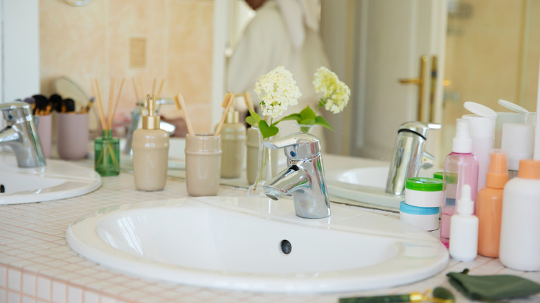 Cluttered bathroom countertop with person walking out of bathroom.