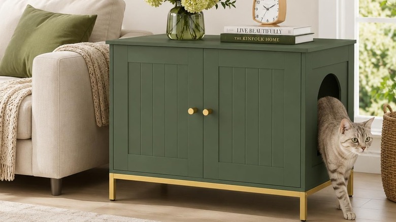 The Homhedy Cat Litter Box Enclosure in green next to a cream couch with a cat coming out of the enclosure's hole