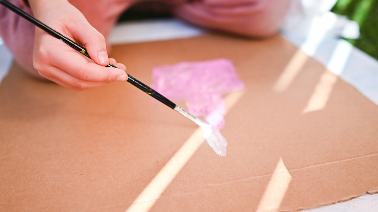 Person's hand painting the bare surface of a sheet of cardboard.