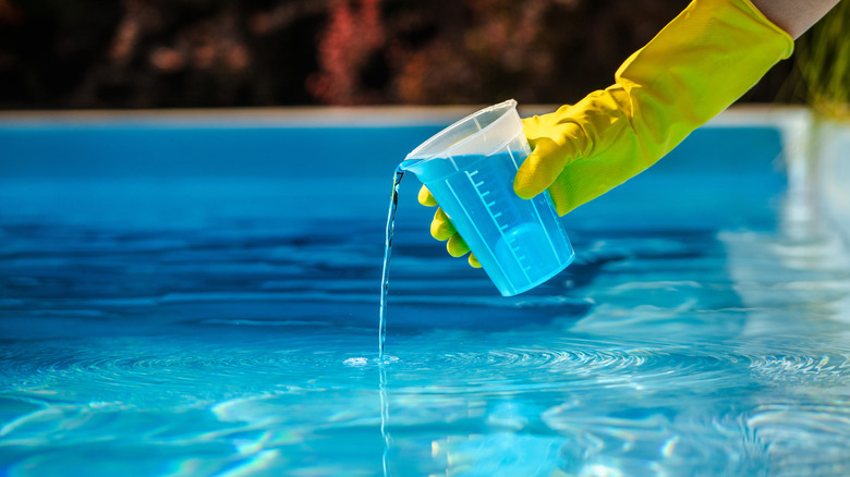 A person wearing a yellow rubber gloves pours a blue liquid chemical into a swimming pool