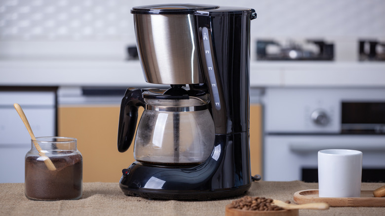 A black drip-coffee maker in a kitchen with ground beans in a jar and a white coffee cup.