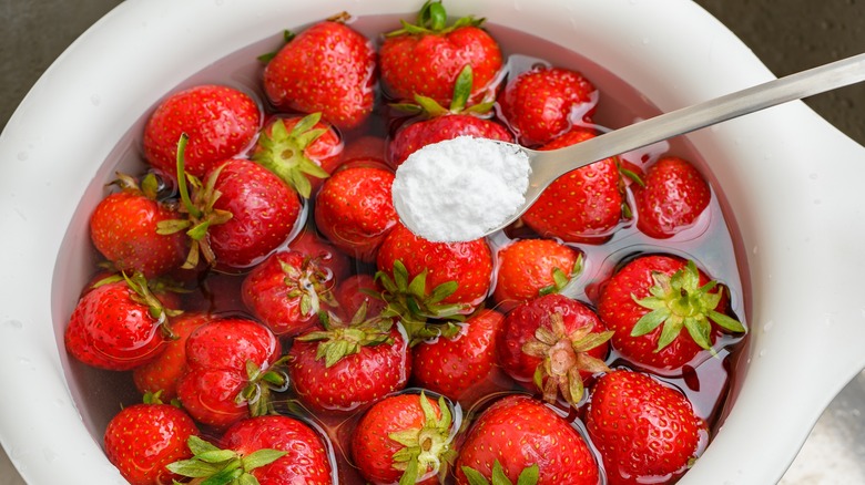 A bowl of strawberries in water with a spoon of baking soda hovering over it.