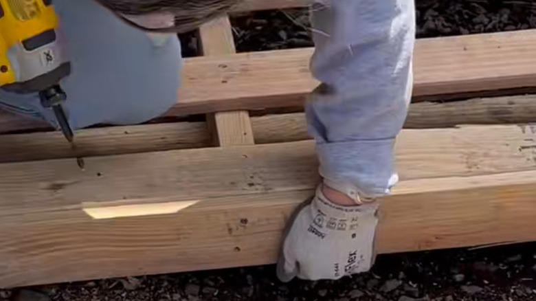 A person  with gloves holding a wood panel onto the end of a wood pallet while holding a drill
