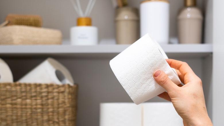 A hand holding a toilet paper roll with more on a shelf in the background.